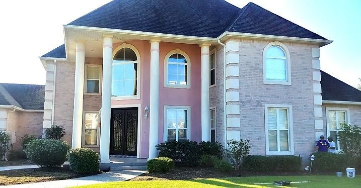 Two-story house with light pink brick exterior, white columns, and a dark roof. A person paints outside.