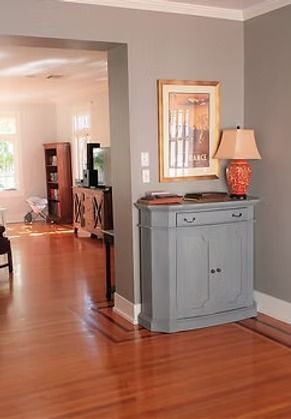 A gray cabinet with a lamp and framed artwork sits in a hallway. Warm hardwood floors and gray walls are visible.