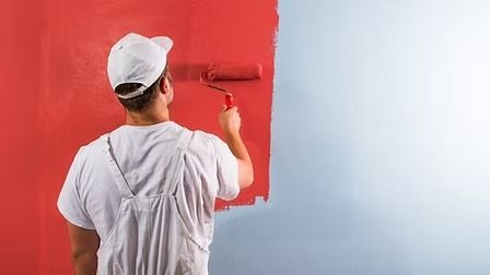 Painter in a white cap and shirt, using a roller to apply red paint to a wall; a section of wall is still blue.