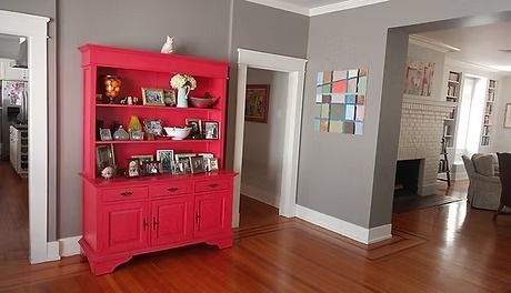 A red hutch filled with items sits in a room with grey walls and hardwood floors. The room has open doorways and a wall with artwork.