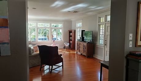 A view into a bright living room with a burgundy armchair, hardwood floors, and large windows.