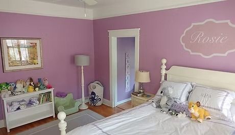 Child's bedroom with lavender walls, white bed, and a custom name decal. Includes a white bookshelf with toys and a gray floor lamp.