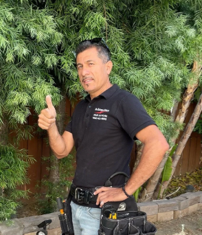 Man in black polo and jeans giving a thumbs-up beside greenery and a wooden fence