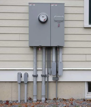 Gray electrical meter and service boxes mounted on a house exterior with conduit pipes beneath.
