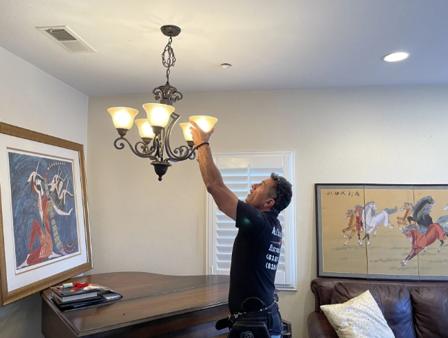 Person reaches up to adjust a chandelier in a living room with framed art and a couch.