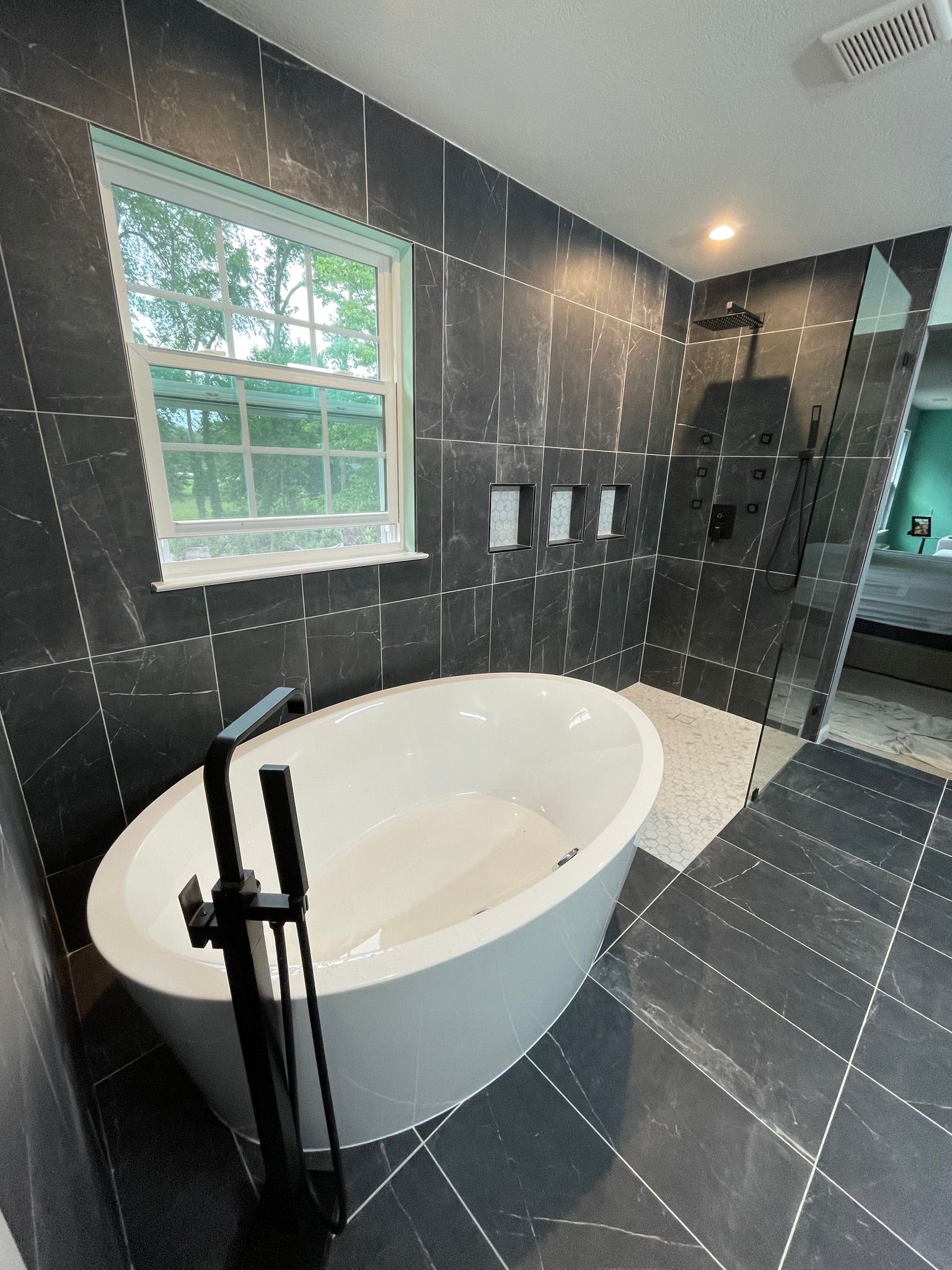 Modern bathroom with a white oval bathtub, black tile, a shower, and a window.
