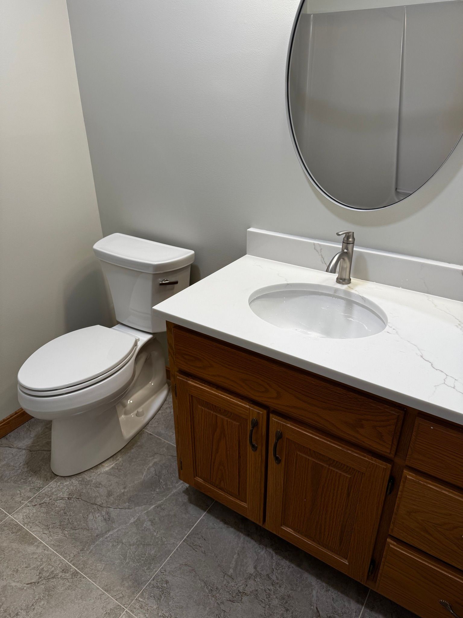 Bathroom with a white toilet, brown vanity, round mirror, and gray walls.