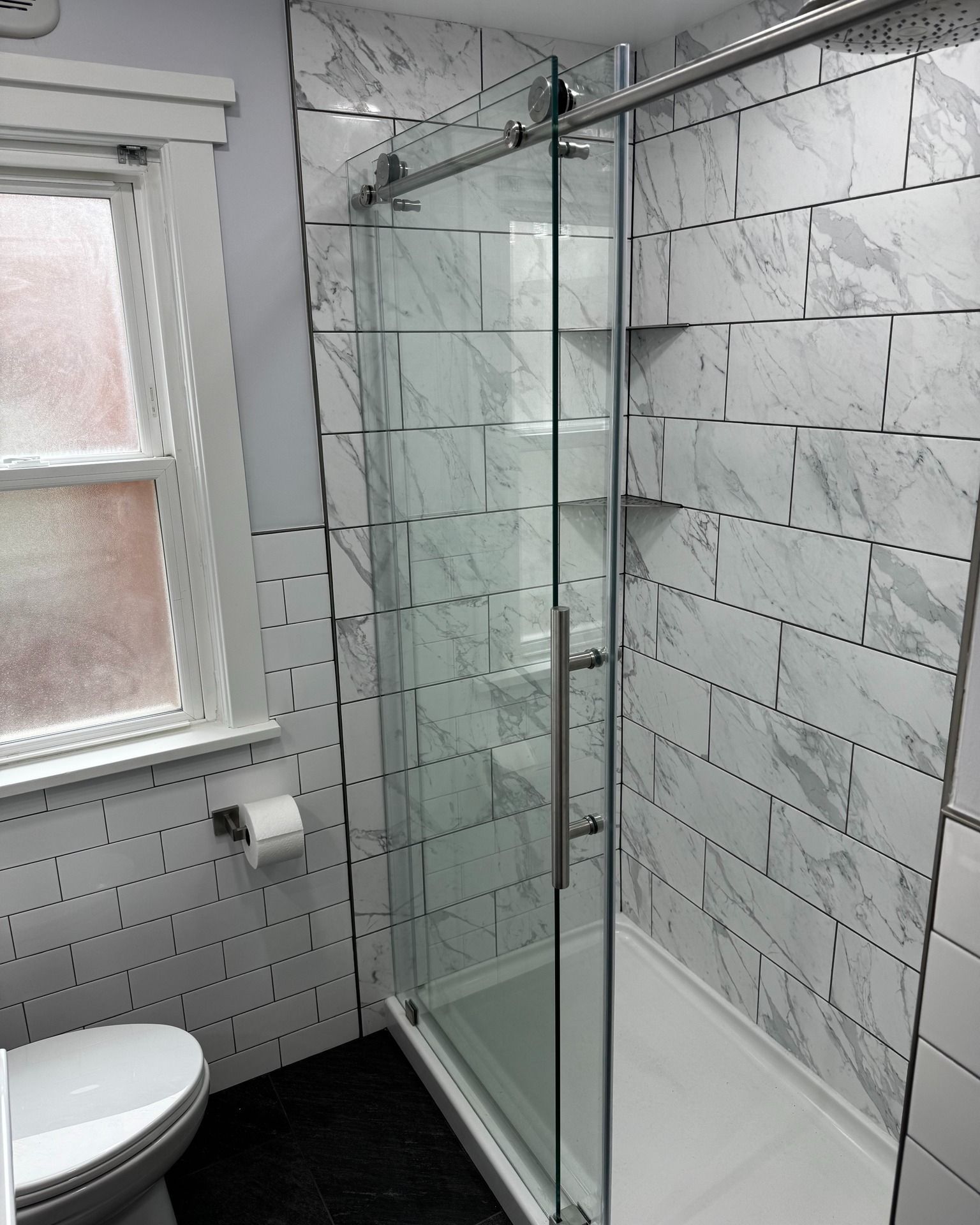 Bathroom with marble-style tiled walls, glass shower with sliding door, white toilet, and dark floor.