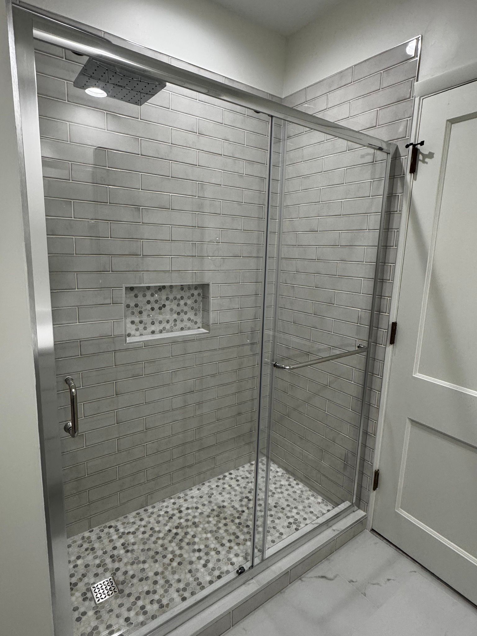 Modern shower with sliding glass doors, gray tiled walls, pebble floor, and a recessed shelf.