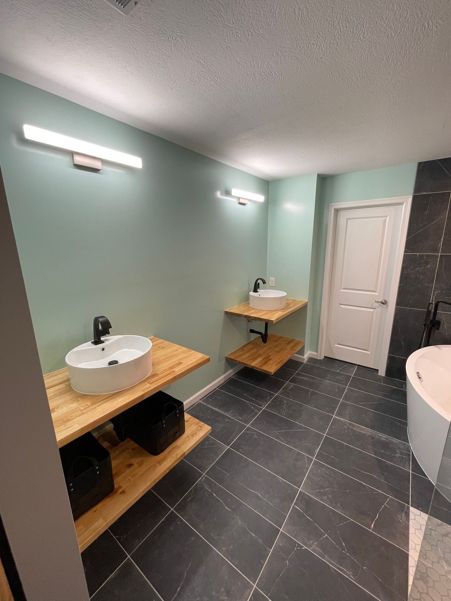 Bathroom with two floating wooden shelves holding white sinks, black fixtures, and black tile floor.