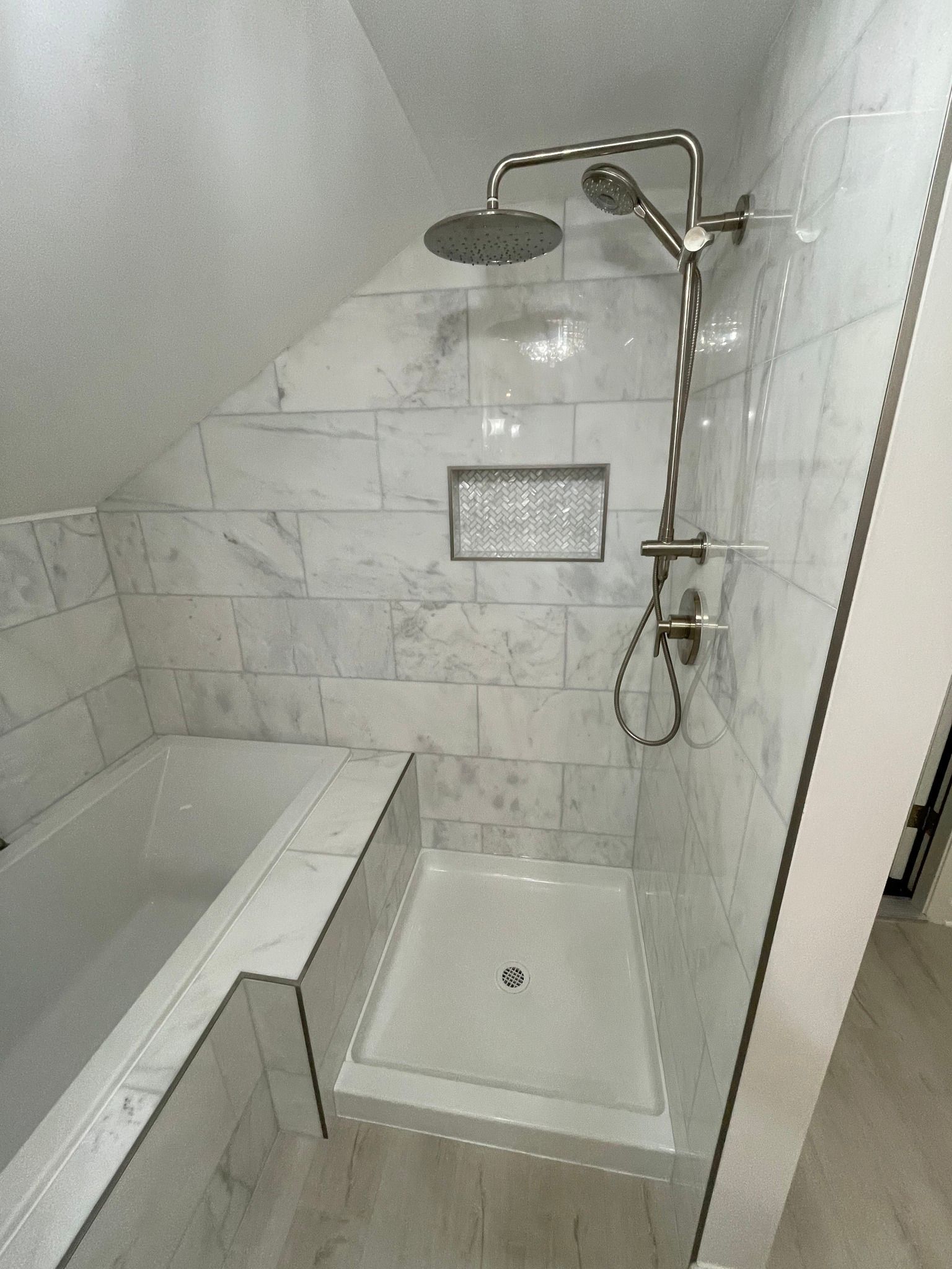 Tiled bathroom with showerhead, handheld shower, and bathtub. White and gray tones, angled ceiling.