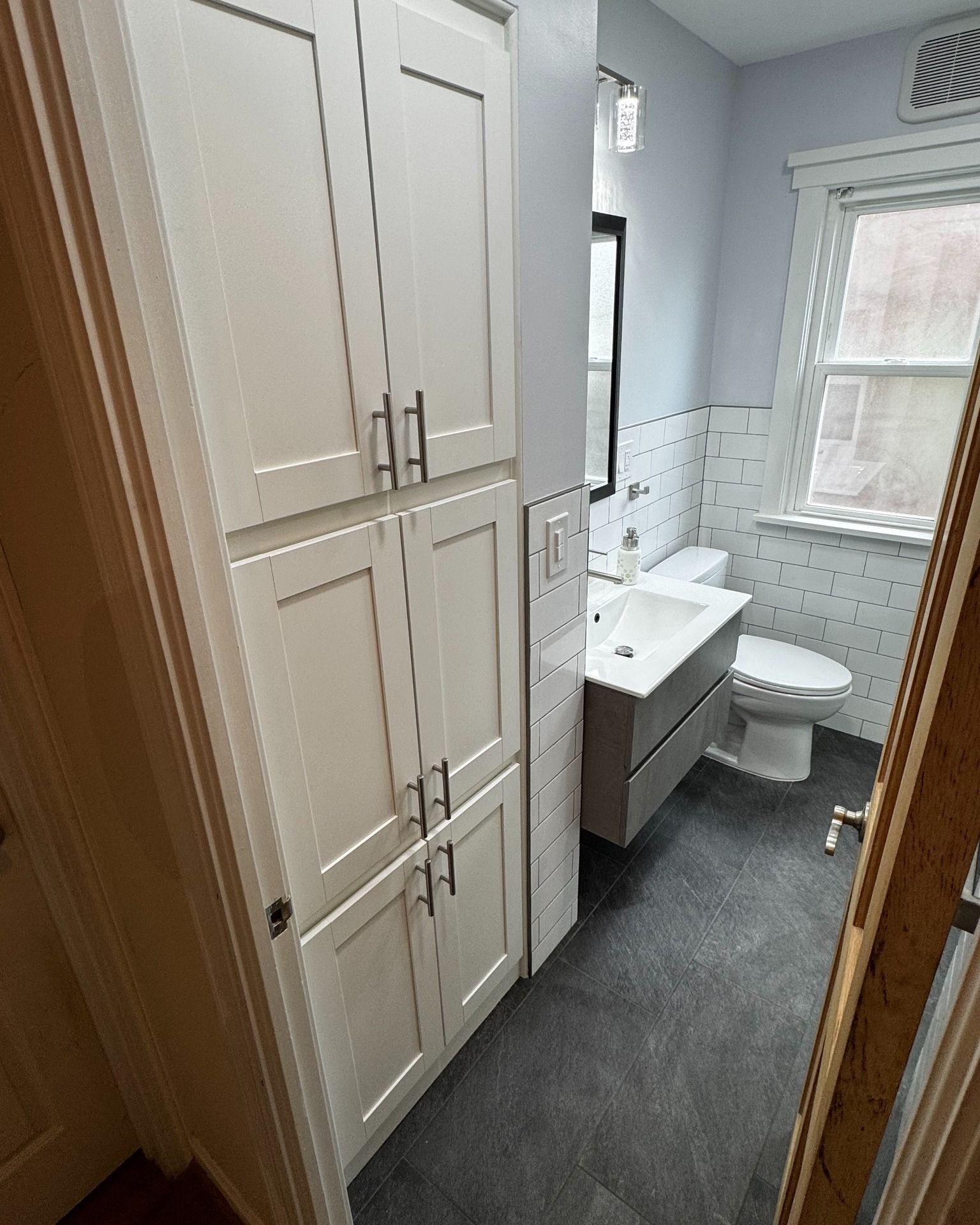 Small, modern bathroom with tall white storage cabinet, vanity, and toilet. Dark floor, light blue walls, and window.
