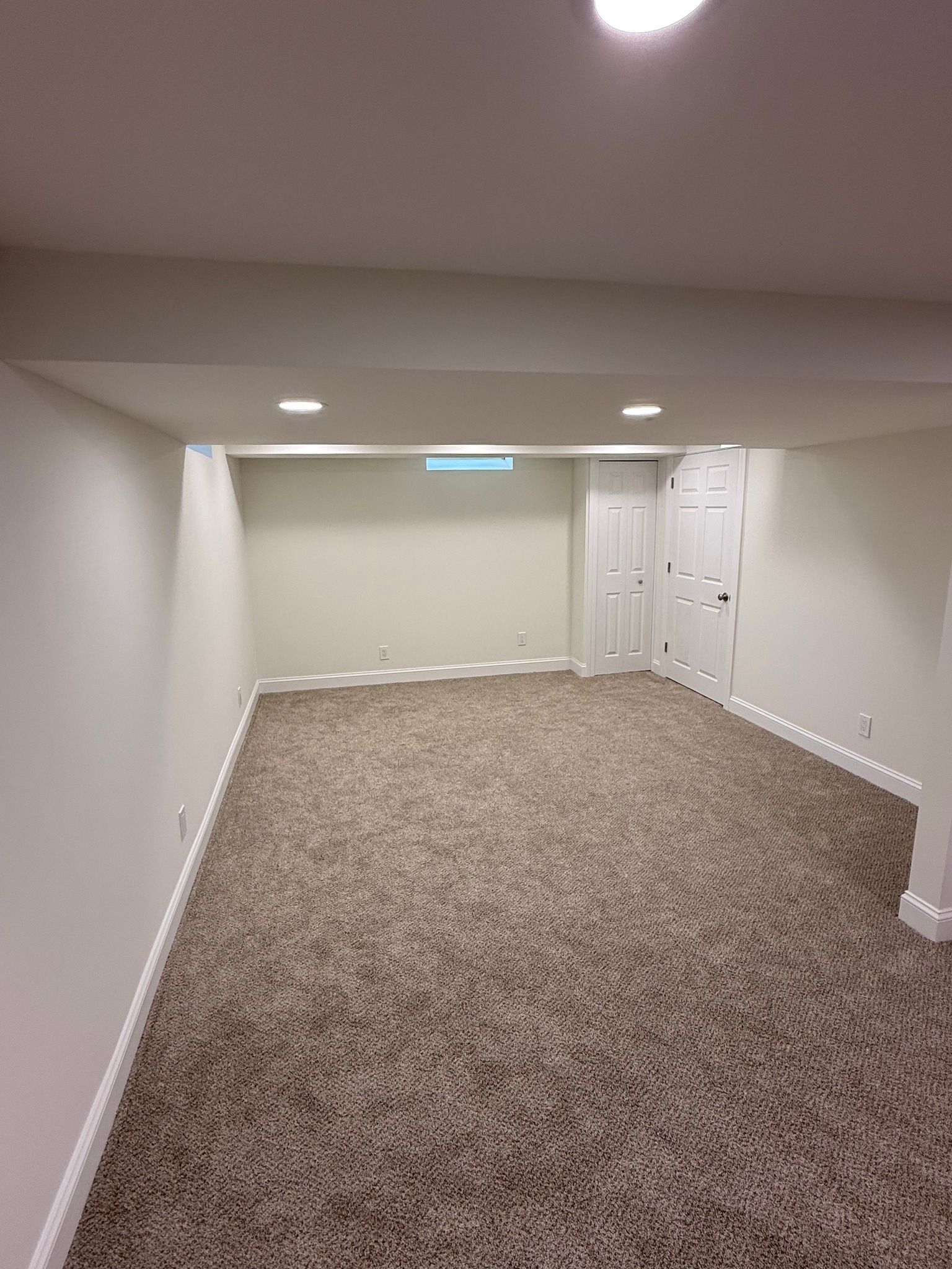 Empty basement room with brown carpet, white walls, recessed lighting, and closed white doors.