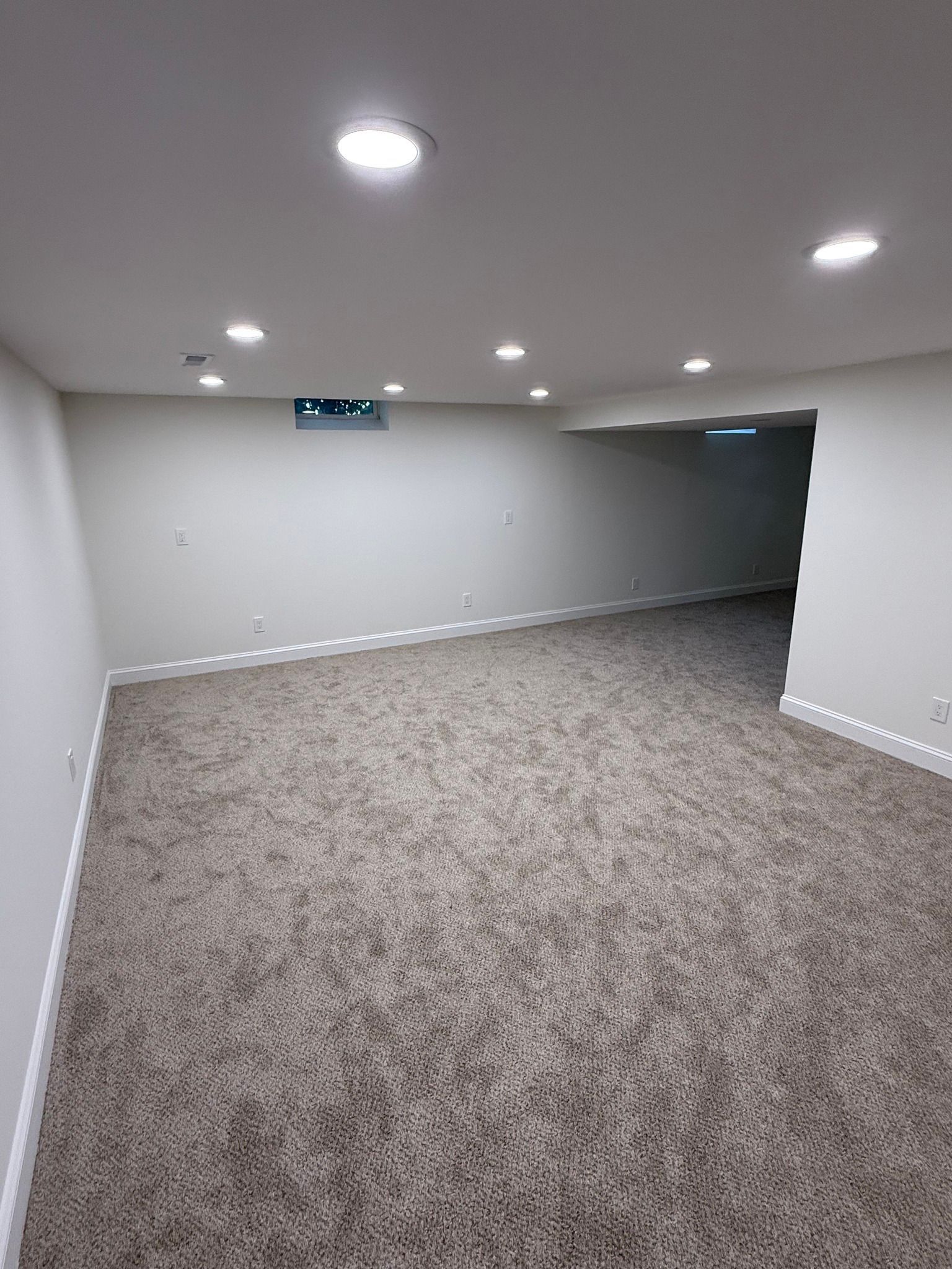 Empty basement room with neutral-colored walls and carpet; recessed lighting, open doorway to another room.