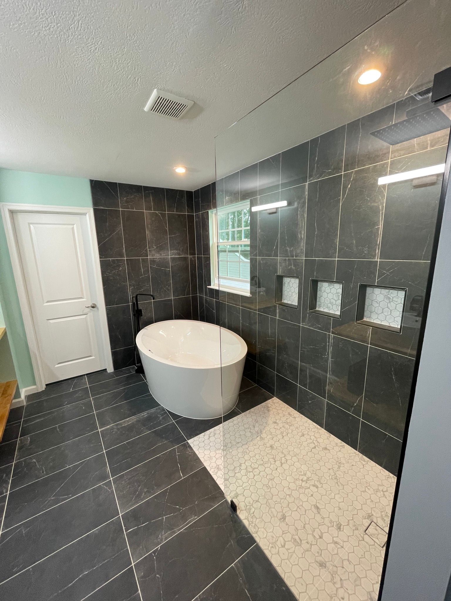 Bathroom with dark tile walls and floor, white tub, shower niche, and small window.