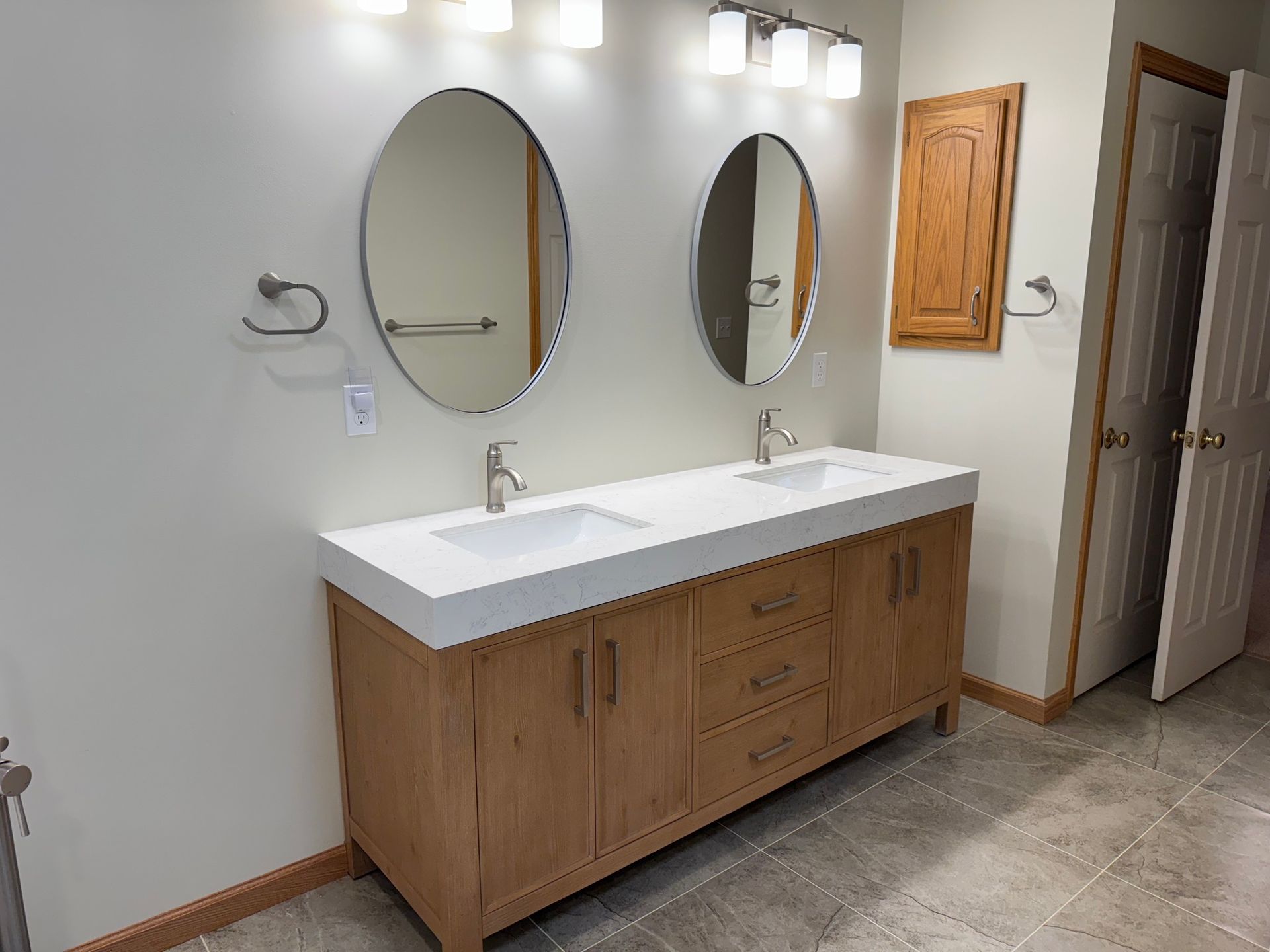 Bathroom with wood vanity, oval mirrors, white countertop, and double sinks.