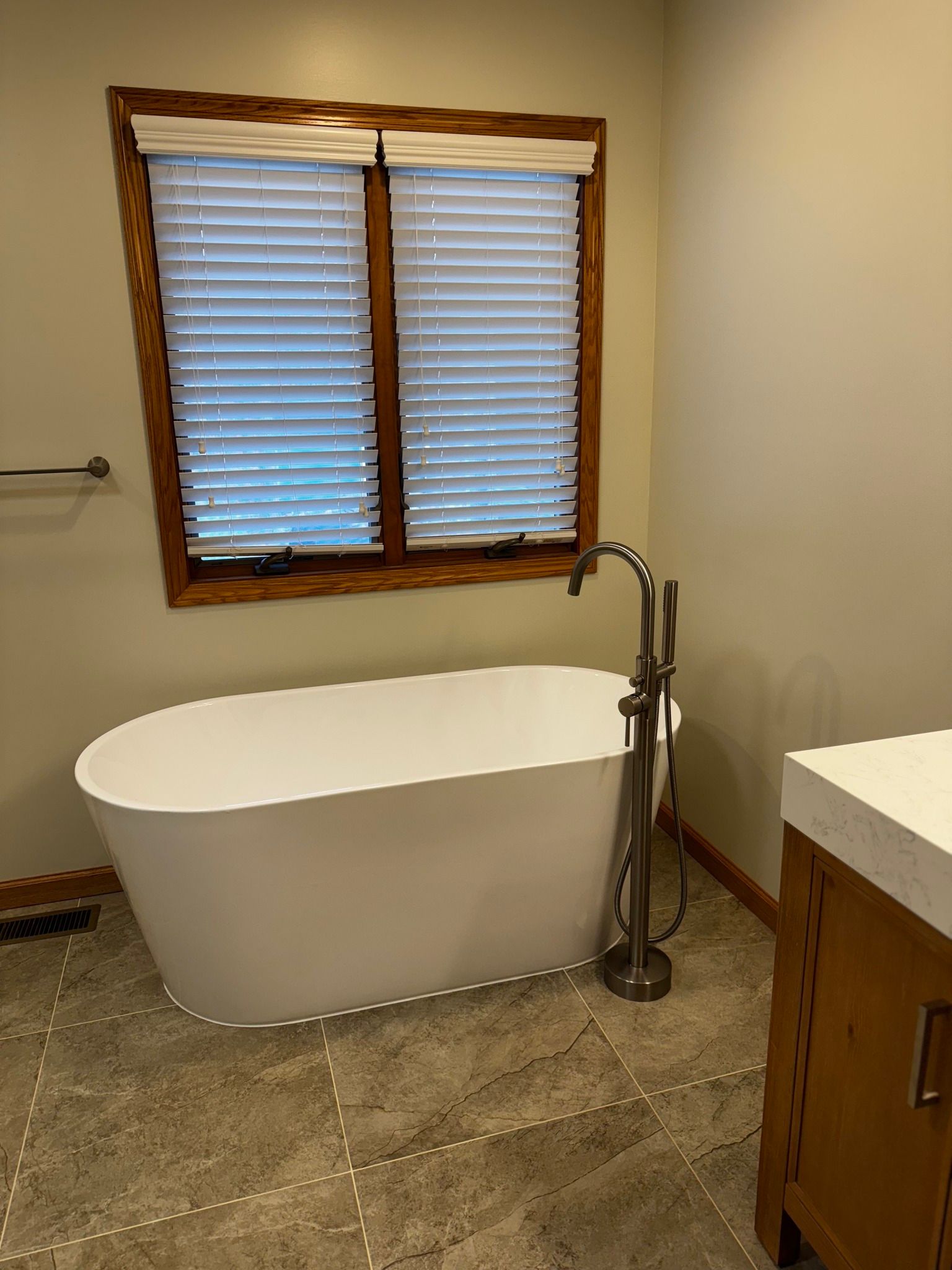 Freestanding white bathtub by window with blinds in bathroom; brown-framed window, wooden cabinet, and gray tile floor.