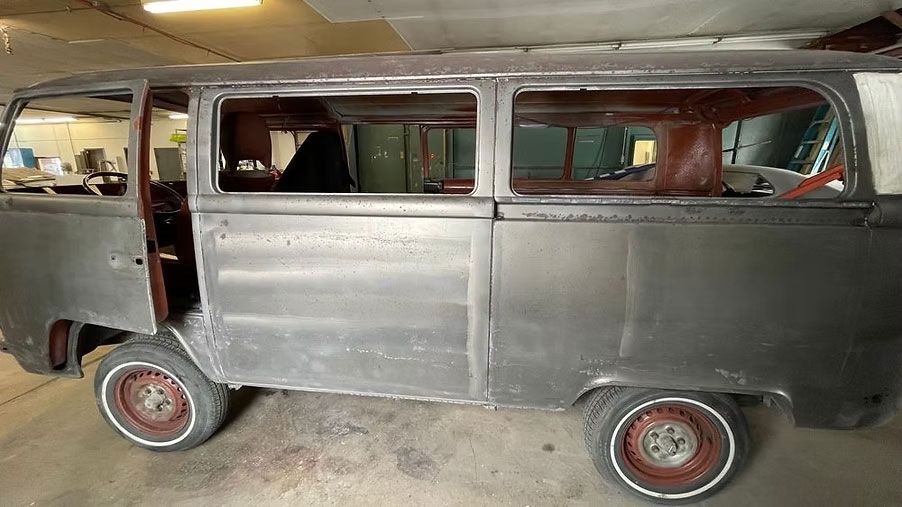 Gray and red stripped-down VW van in a workshop; doors and windows open, wheels attached.
