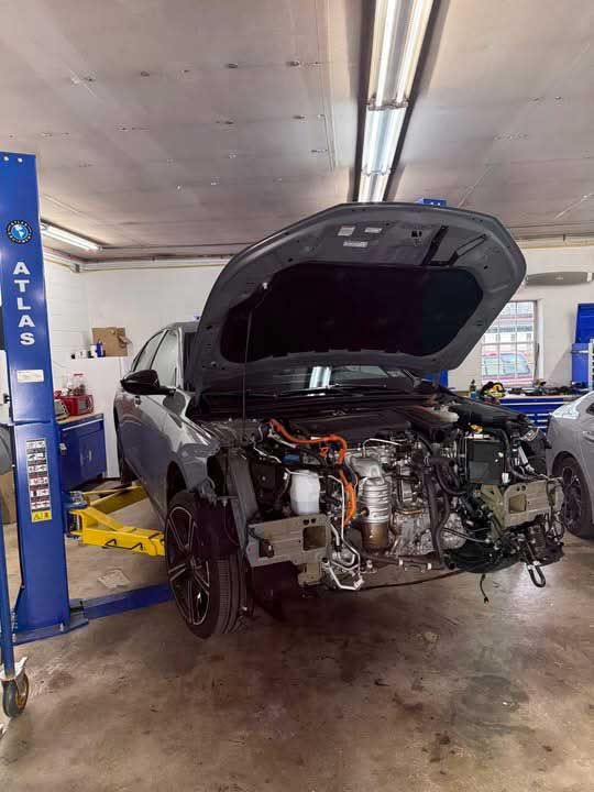 Car on a lift with hood open in a repair shop; front bumper removed.
