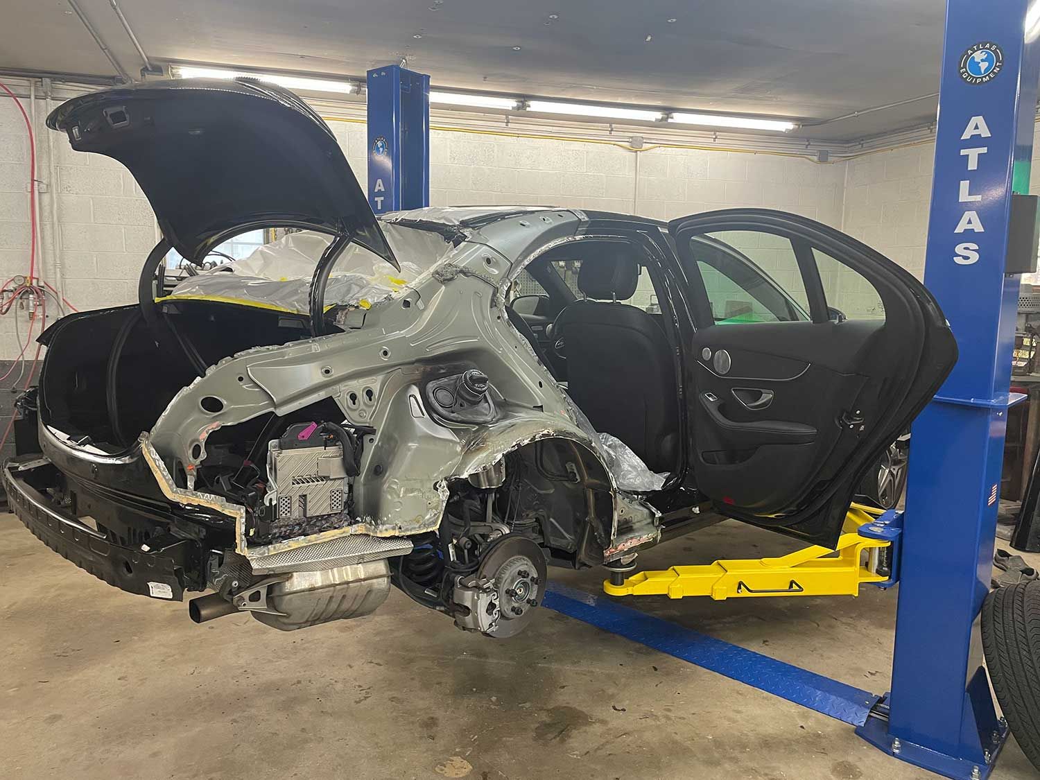 A black car body on a lift in a repair shop; rear end is disassembled, trunk open.