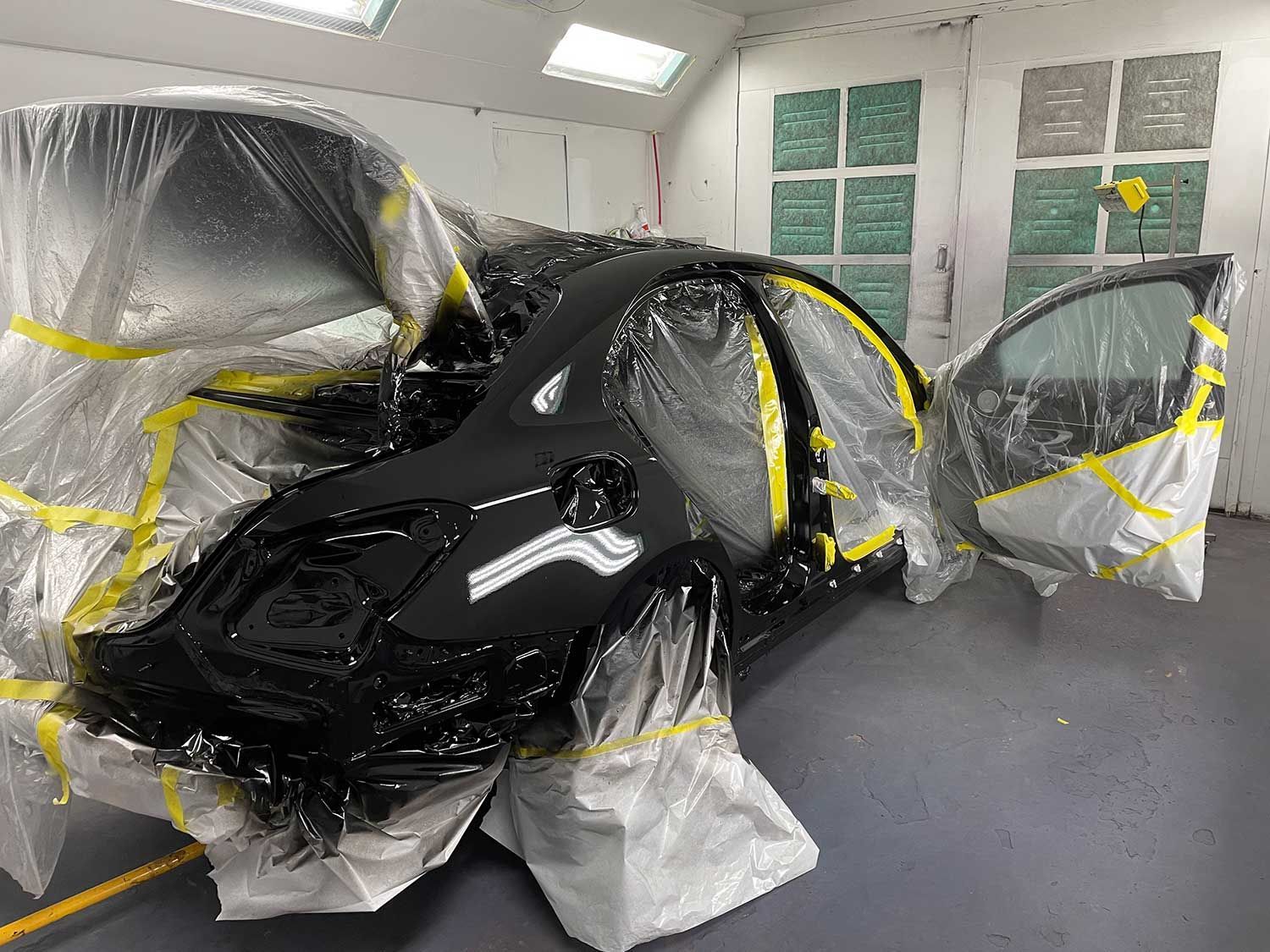 Black car in a paint booth, partially covered with plastic and yellow tape for painting.