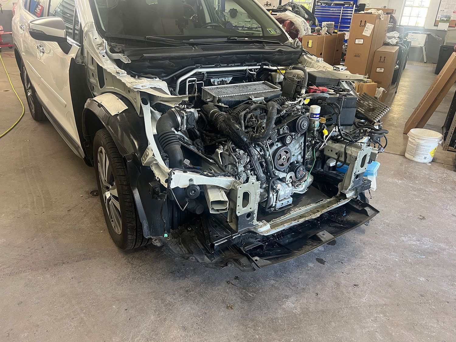 White car with damaged front, engine exposed in a repair shop setting.