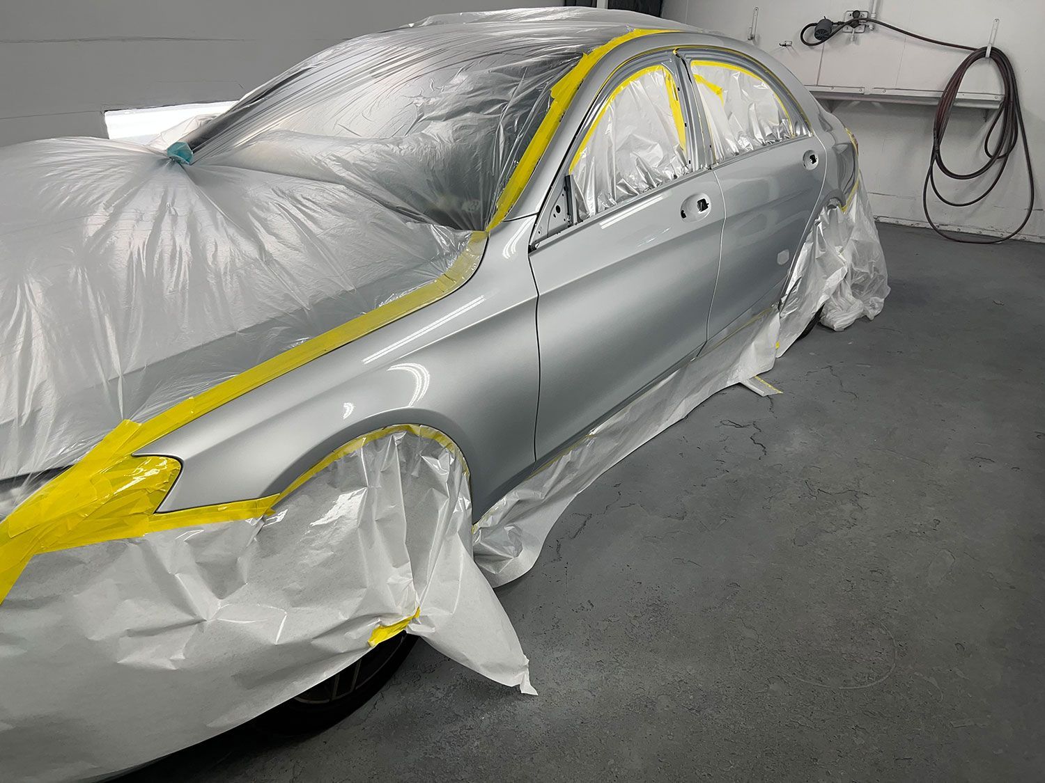 Silver car masked with plastic and yellow tape, ready for painting in a workshop.