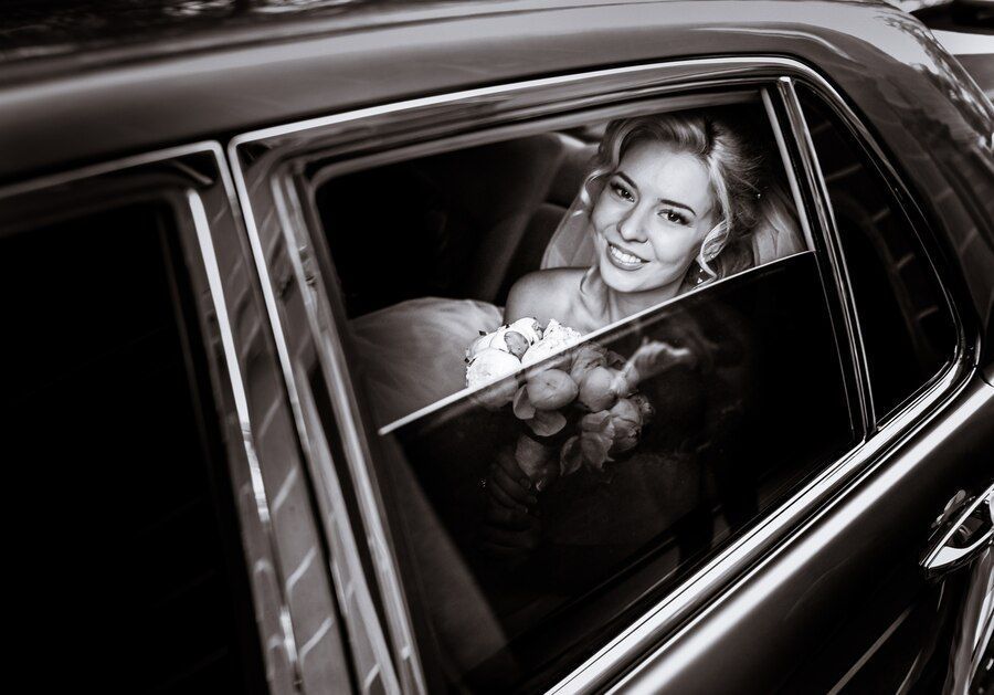 Bride in a car smiles, holding bouquet. Black and white photo.