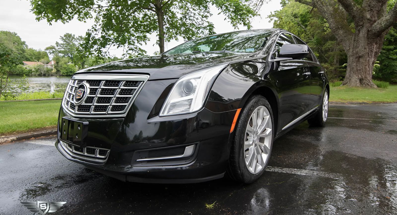 Black Cadillac XTS parked on a wet road, with a pond and tree in the background.