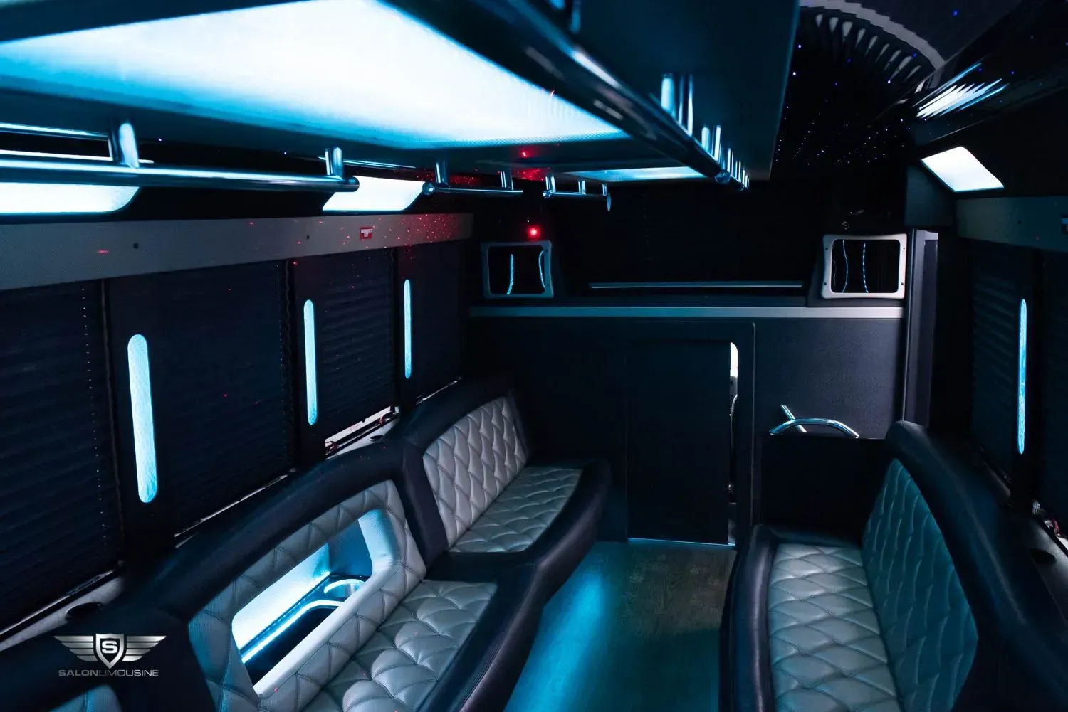 Interior of a black party bus with teal accent lighting, quilted gray leather seating, and a bar area.