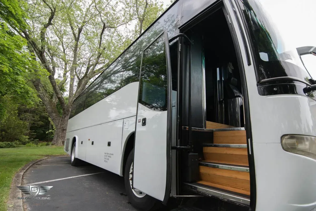 White tour bus with open door and steps on a paved area, with trees in the background.
