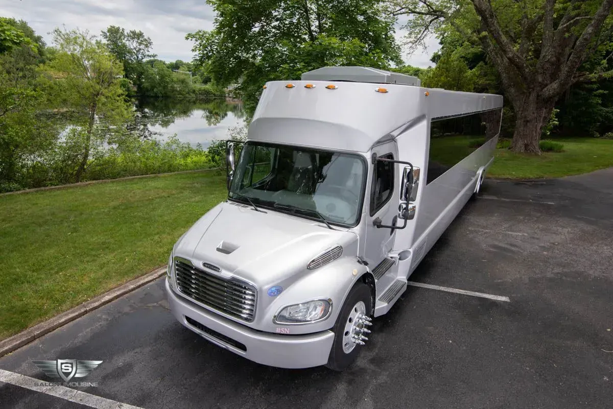 Silver party bus parked on asphalt next to grass and a body of water with trees.