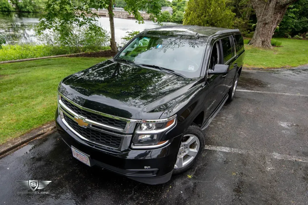 Black Chevrolet Suburban SUV parked on wet asphalt next to green grass and water.