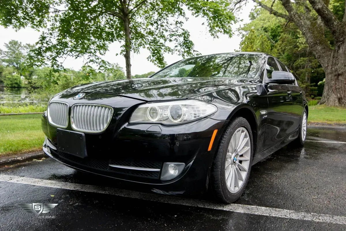 Black BMW sedan parked on wet pavement in front of trees and a body of water.