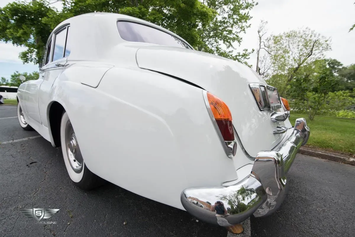 White vintage Rolls-Royce car, rear view. Chrome accents, white tires, parked on asphalt.