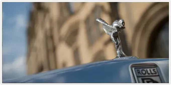 Silver Rolls Royce hood ornament, a Spirit of Ecstasy statue, against a blurred building background.