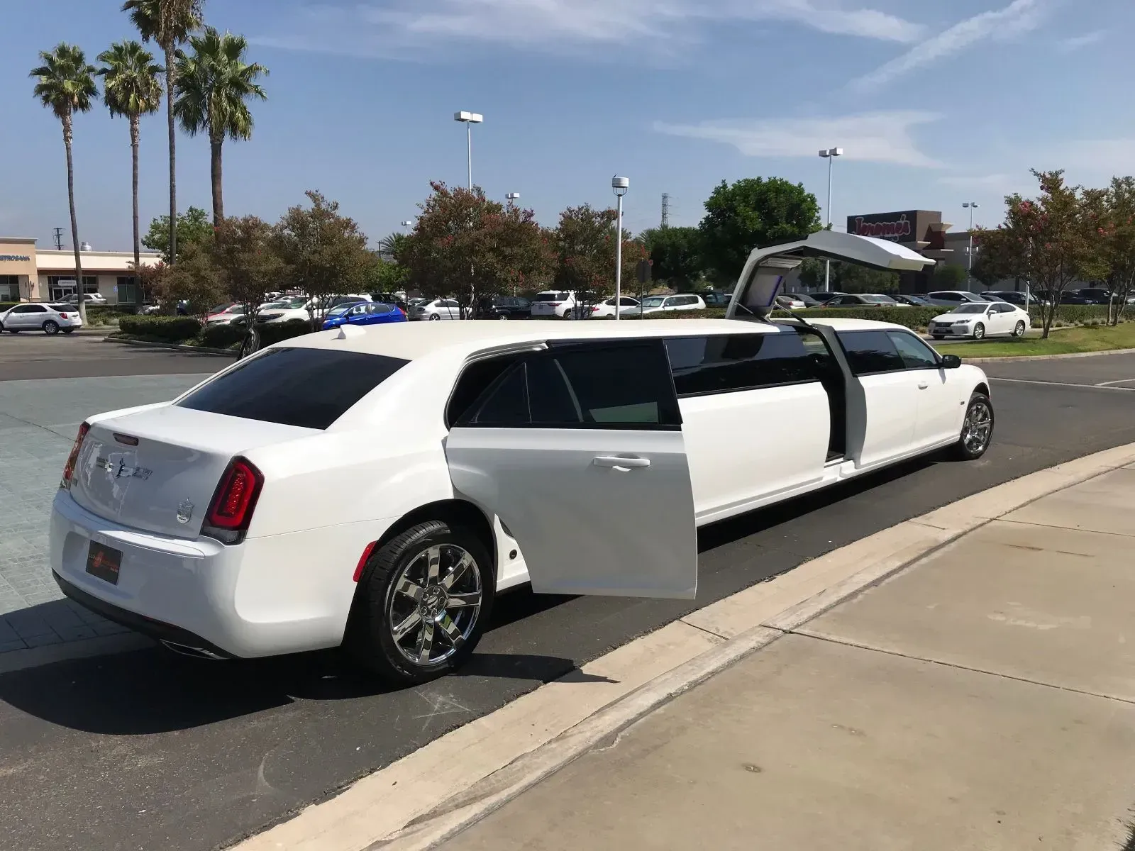 White limousine with open doors parked on a paved area, trees and businesses in the background on a sunny day.