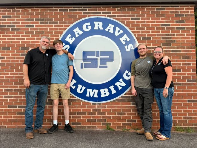 Four people standing in front of a Seagraves Plumbing sign on a brick wall