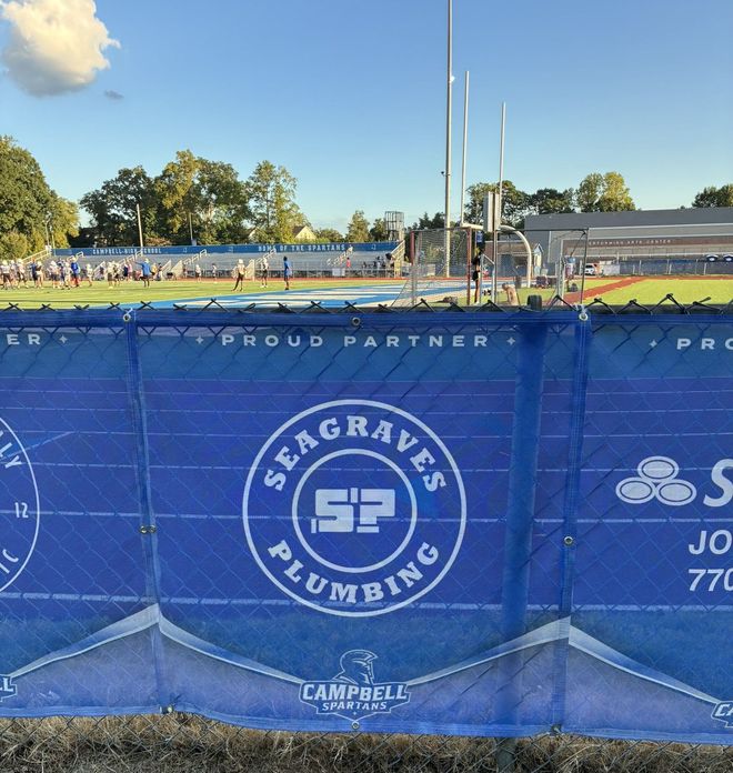 Blue sponsor fence at a sports field with “Seagraves Plumbing” logo and a stadium in the background