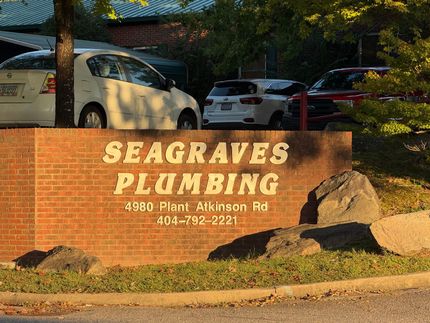 Brick sign for Seagraves Plumbing with address and phone number, in front of parked cars and trees.