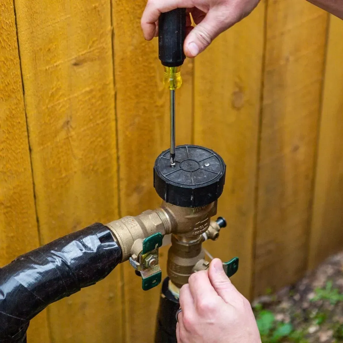 Hands using a screwdriver to adjust a brass outdoor hose valve against a wooden fence.