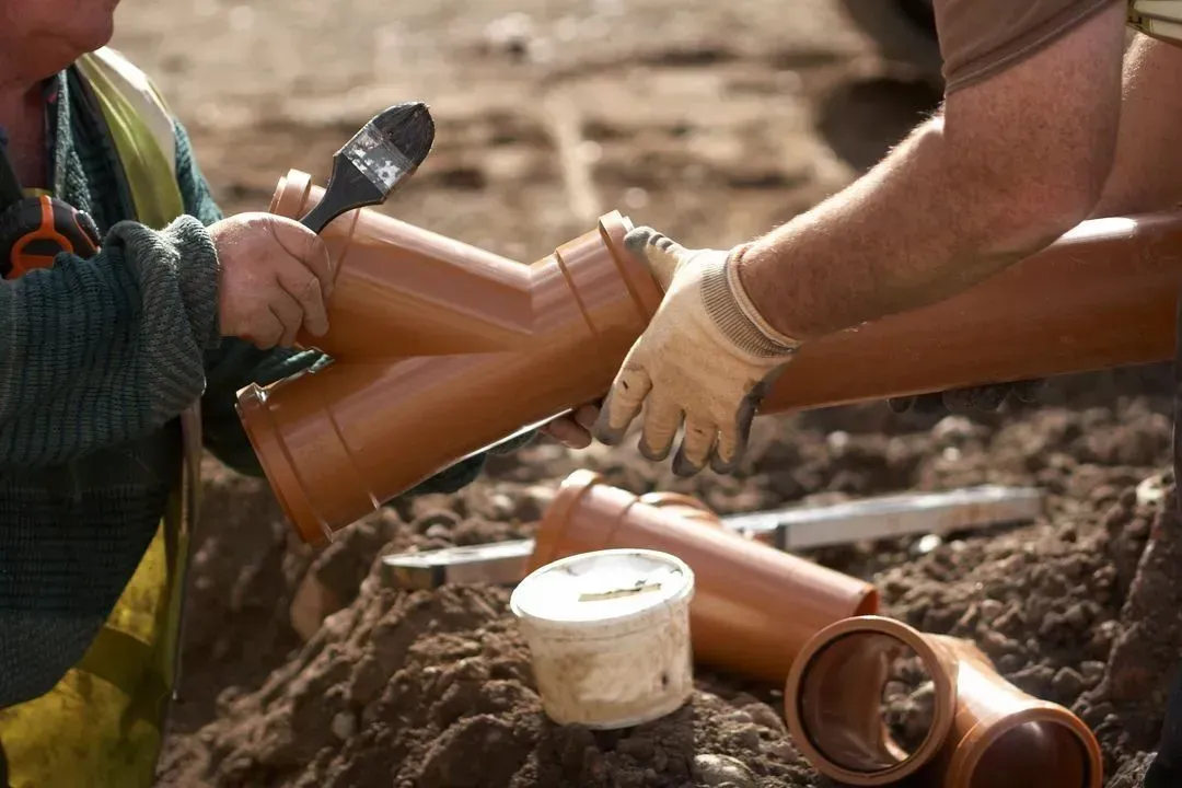 Two workers handle brown PVC pipes on a dirt construction site, with tools and fittings nearby.