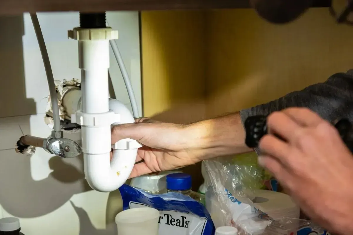 Hand reaching under a sink to tighten a white PVC drain pipe; cleaning supplies nearby.