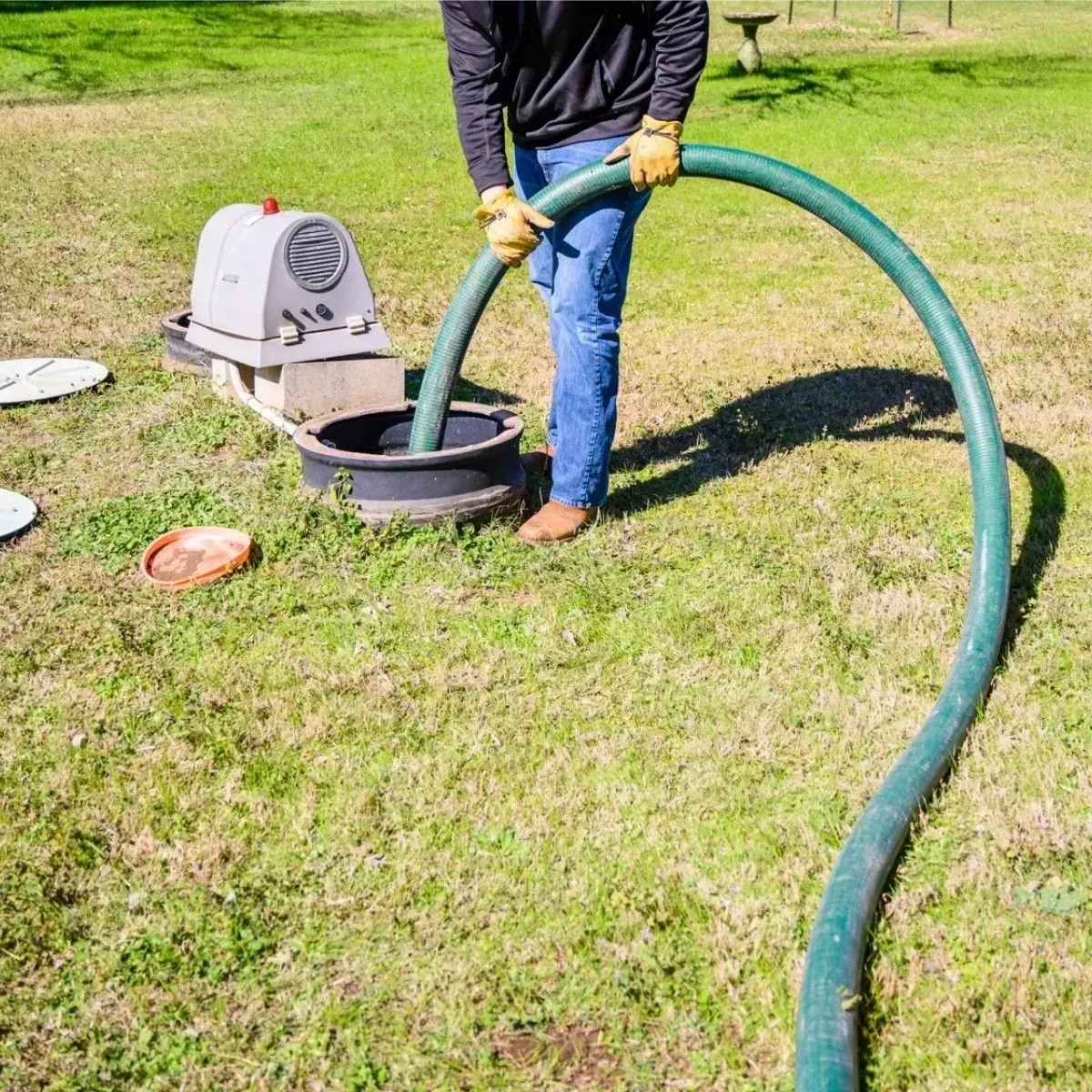 Person using a green hose to fill a black bucket beside a small machine on a grassy lawn