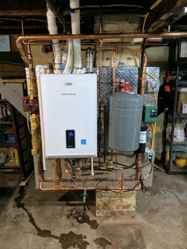 A white water heater is sitting in a basement next to copper pipes.