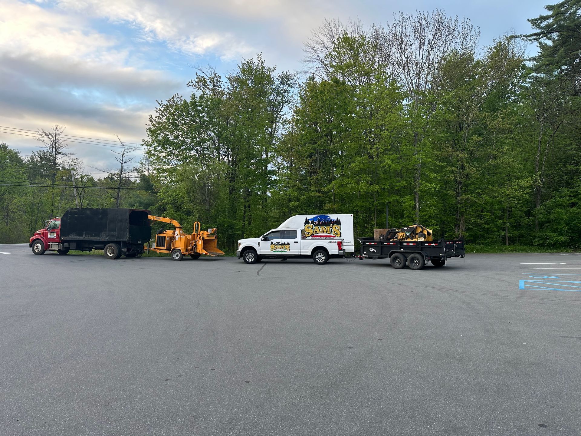 Tow truck hauling a white service truck and orange equipment on a lot beside trees