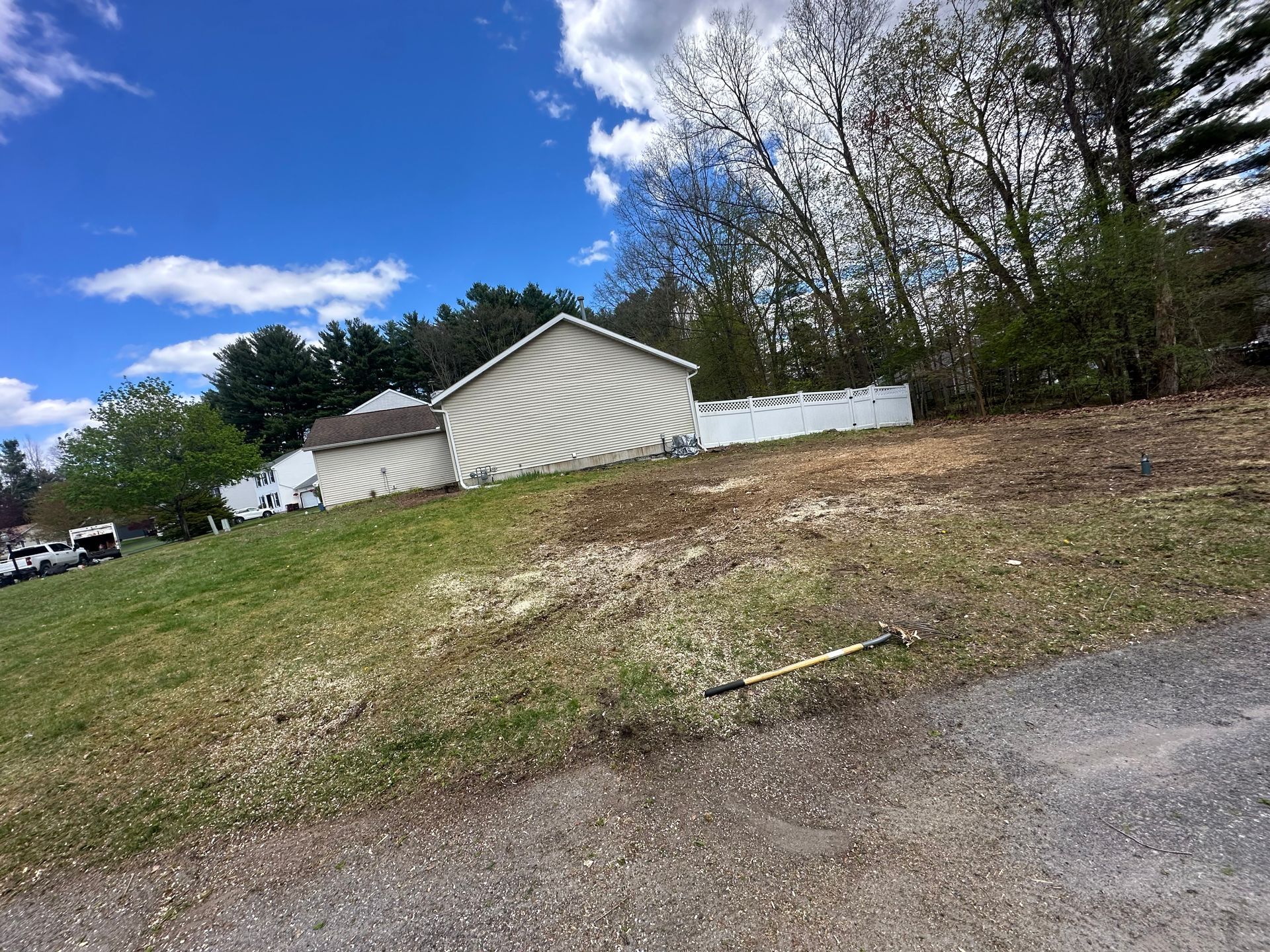 Grassy vacant lot beside a gravel road, with a white building and trees under a partly cloudy sky