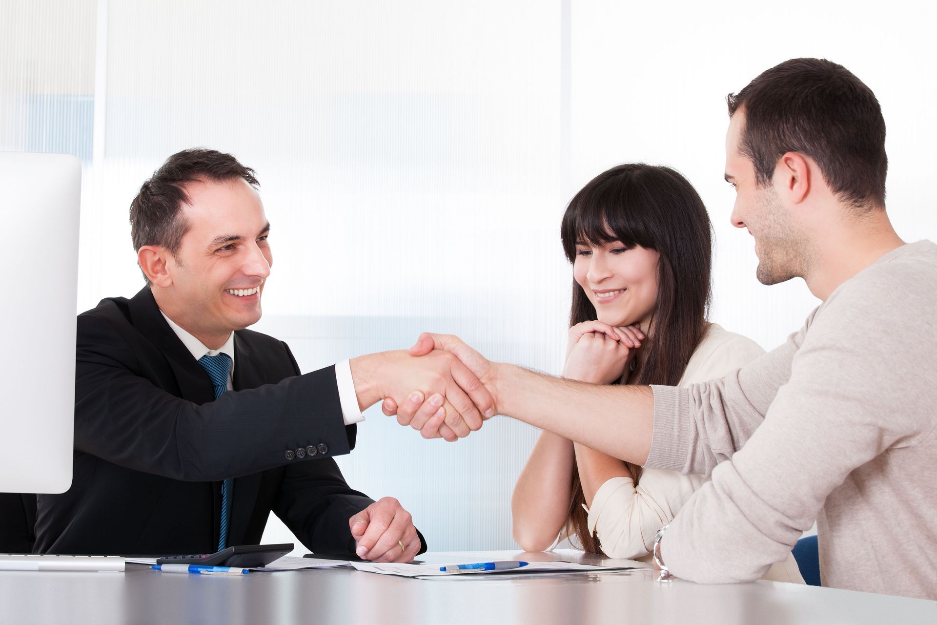 A man in a suit shakes hands with another man at a desk, as a woman smiles nearby; possibly a business deal.