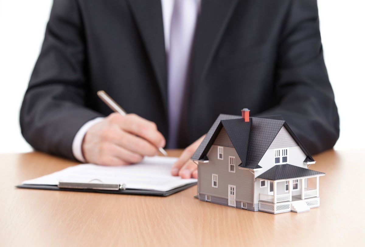 A person in a suit writing on a clipboard, with a model house on the desk.