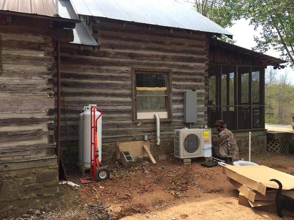 A man is working on HVAC on the side of a log cabin.